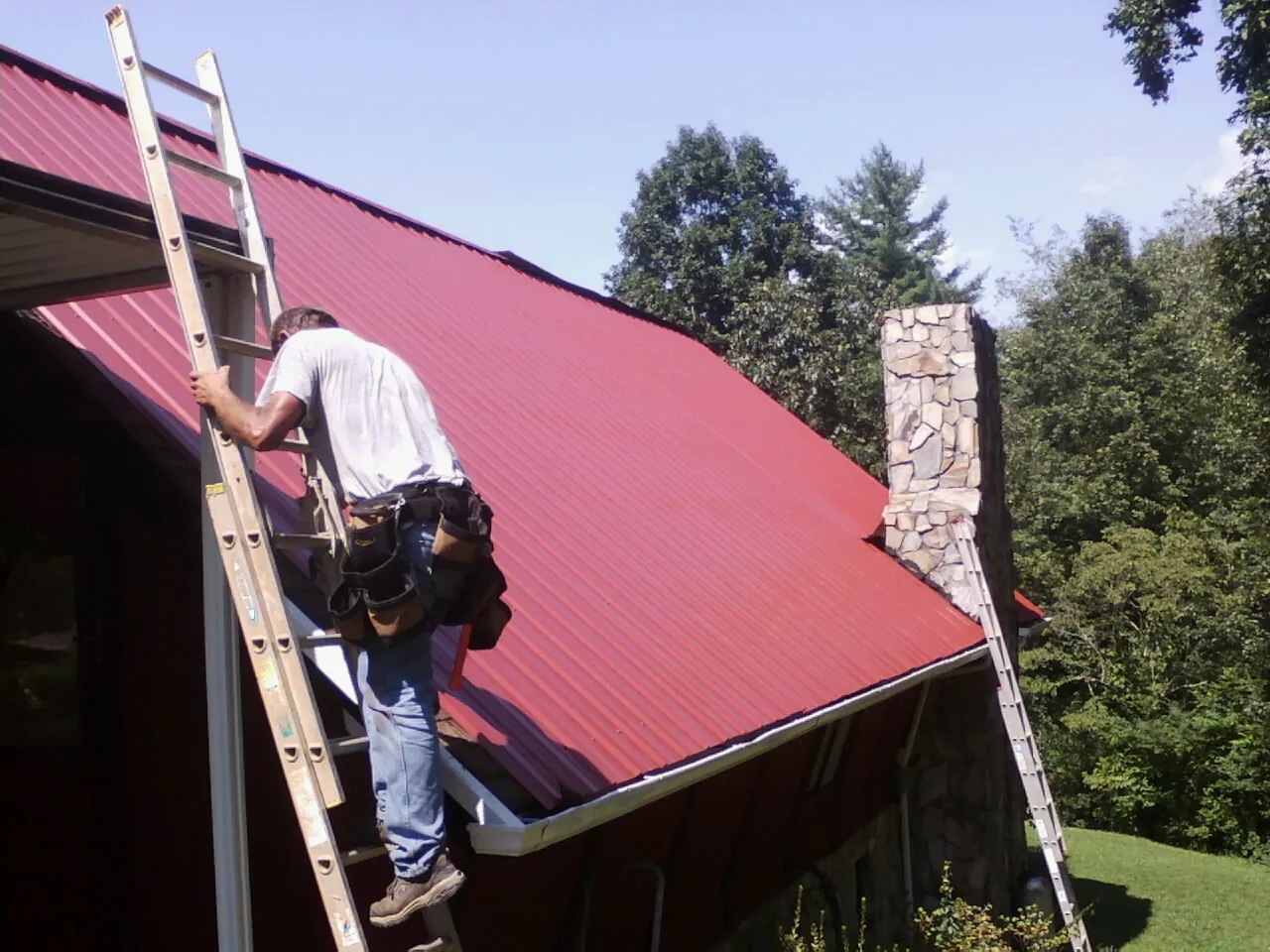 Roofer on a ladder beside a red metal roof and stone chimney — Soffit & Fascia Repair by Roofing Brothers in Jericho